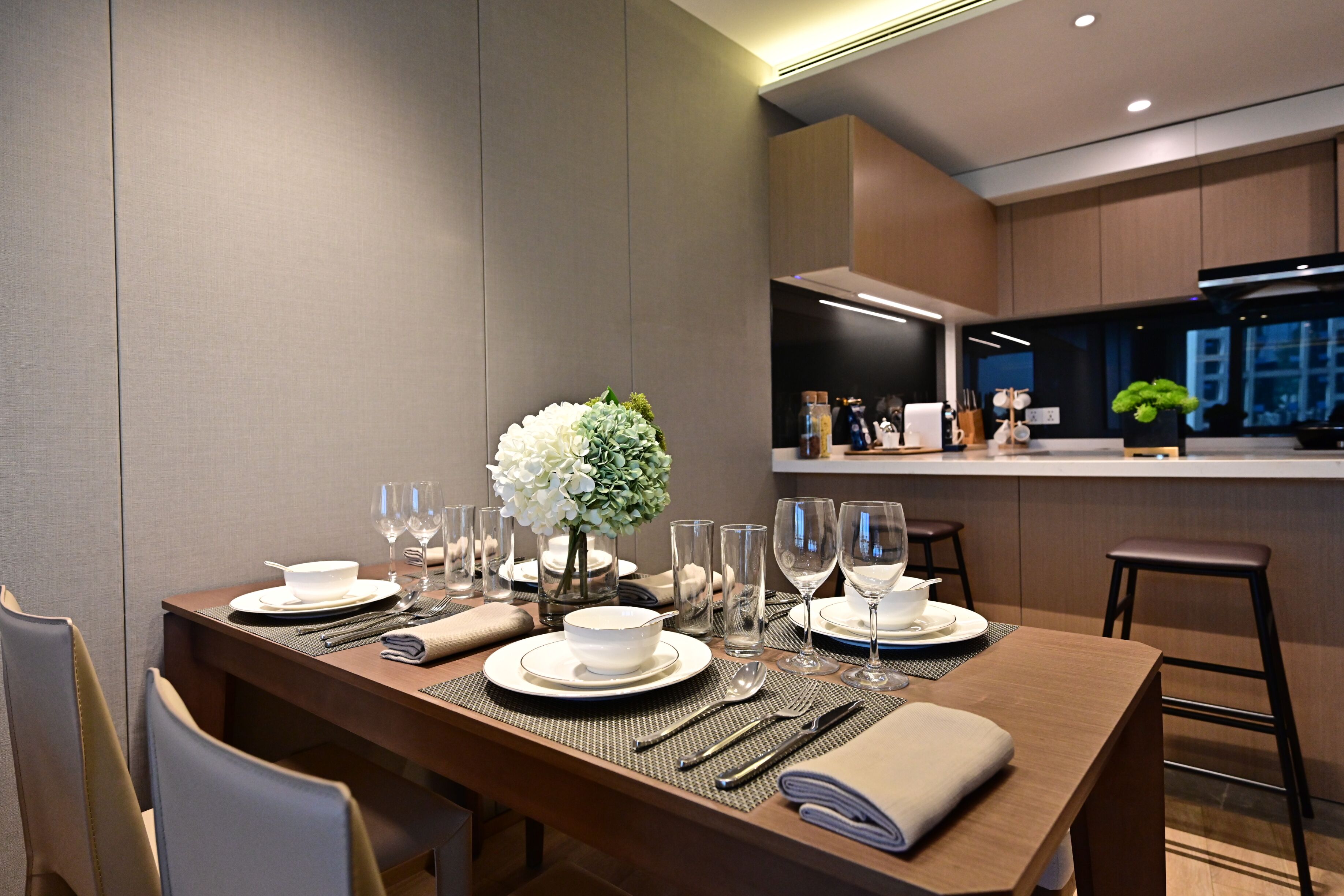 Un comedor moderno con una mesa larga dispuesta para cuatro personas, incluyendo platos, cubiertos y un jarrón con flores blancas.