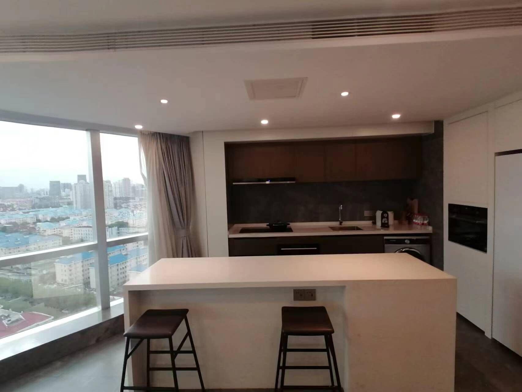 A modern kitchen with a central island, two bar stools, and a view of the city through large windows.