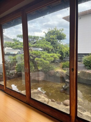 Blick durch ein schmiedeeisernes Fenster auf einen traditionellen japanischen Garten mit Teich, Felsen und verschiedenen Bäumen.