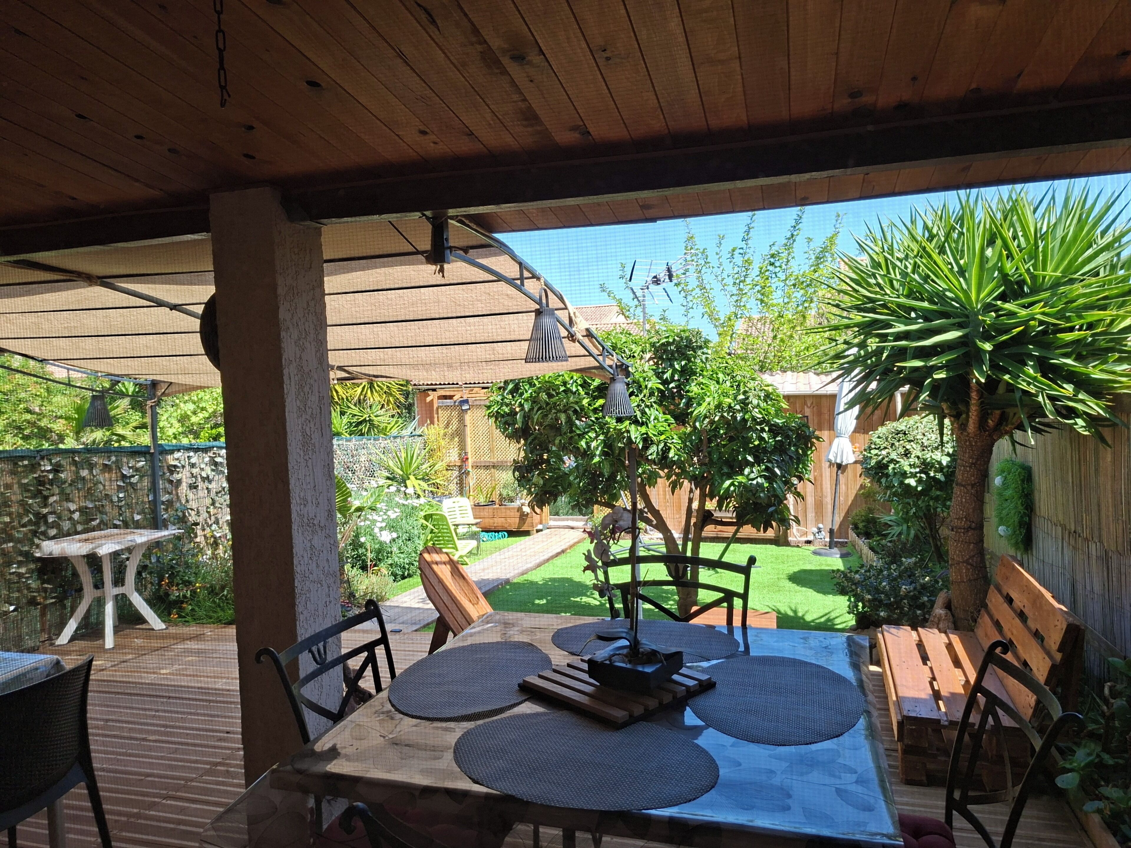 Une terrasse extérieure avec une table et des chaises en bois, entourée de verdure et offrant une vue sur un jardin.
