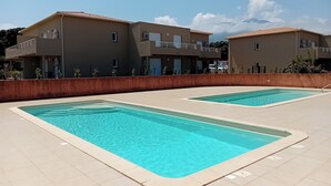 Duas piscinas retangulares com água azul cristalina, rodeadas por um deck revestido de azulejos, e adjacentes a um edifício com varandas.