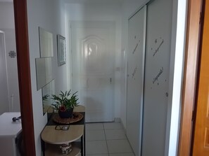 Ein Flur mit einer weißen Tür, einem Spiegel, einem kleinen Tisch mit Pflanze und einer Wand mit dekorativem Text.