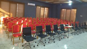 Un salón de conferencias con filas de sillas rojas y negras, techo de madera y ventanas con persianas.
