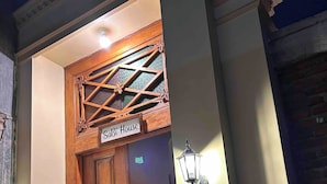 Een houten deur met daar boven een bord waarop 'Sabi House' staat, een decoratief houten plafond en twee wandlampen.