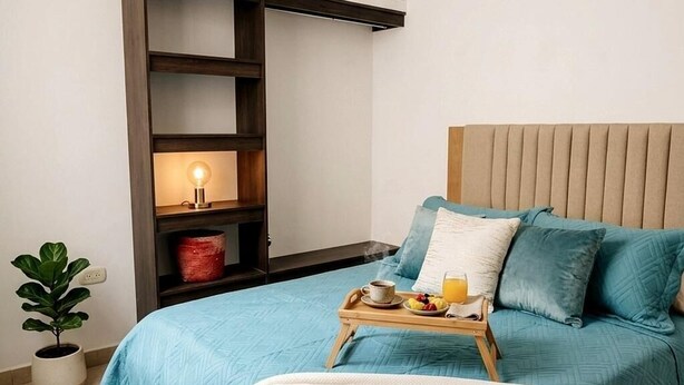 Un lit bien fait, recouvert d’une couvre-lit bleue, une table de chevet avec un plateau de petit-déjeuner, une lampe de chevet et une plante en pot.