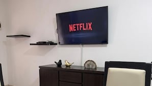 Une pièce avec un téléviseur fixé au mur qui affiche le logo « NETFLIX », un meuble en bois foncé agrémenté d’objets décoratifs et un fauteuil avec un détail rouge.