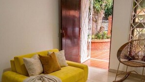 Un canapé jaune agrémenté d’un plaid et de coussins, un fauteuil à bascule en osier et une porte en bois donnant sur un jardin.