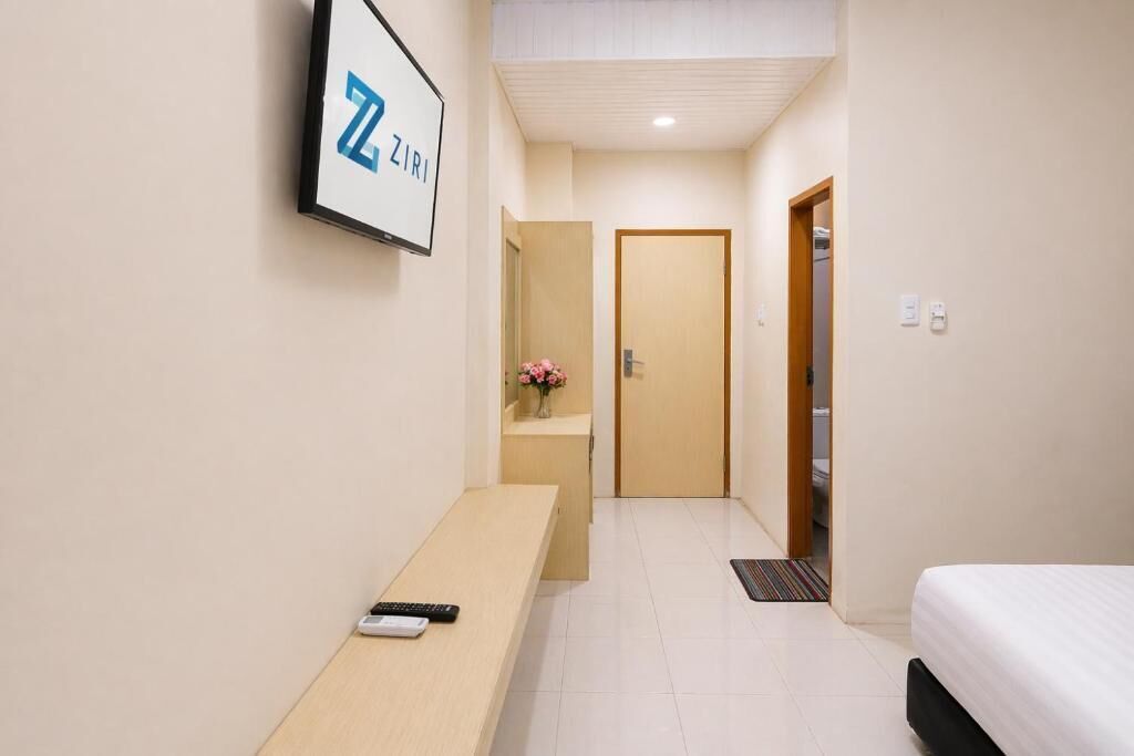 A hotel room with a bed, a desk with a remote control, a framed sign on the wall, and a door leading to another room.