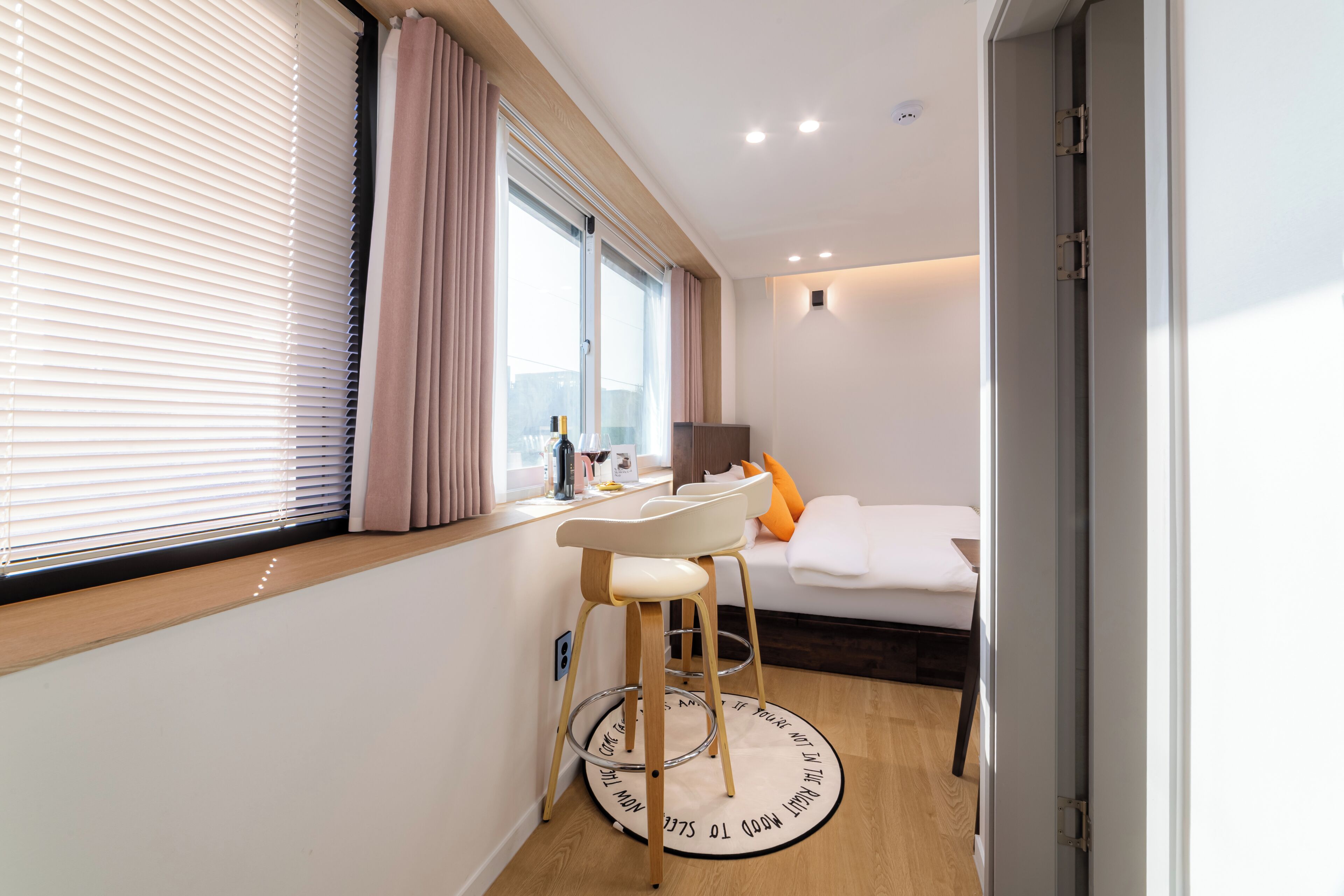 A modern bedroom with a bed, a desk with a chair, a window with blinds, and a door.