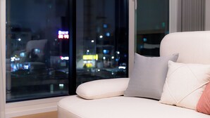 Una sala de estar moderna con un sofá, almohadas y un ventanal con vistas a un paisaje urbano nocturno.