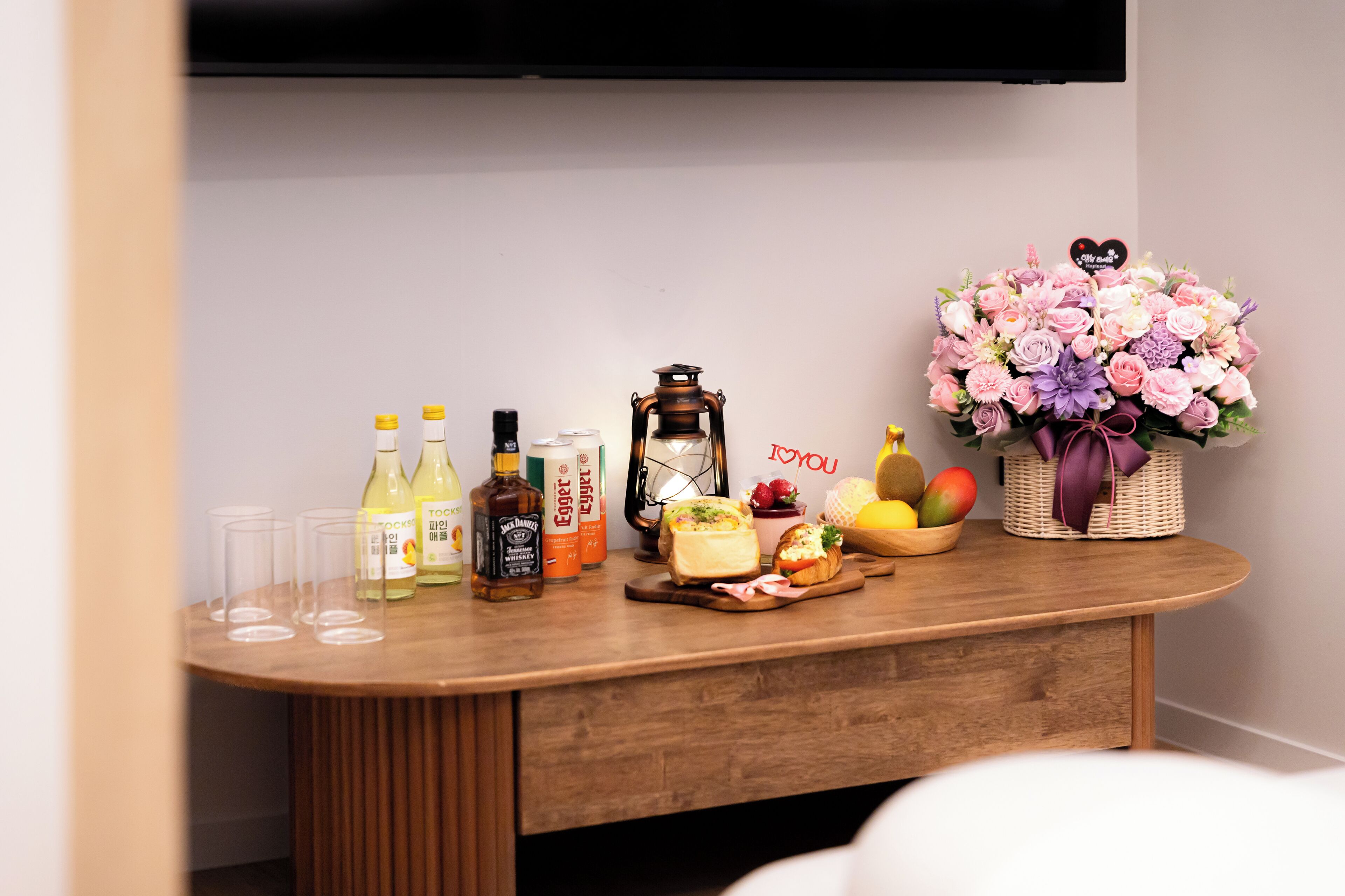 A wooden table with bottles, glasses, a lantern, a fruit basket, and a bouquet of flowers.