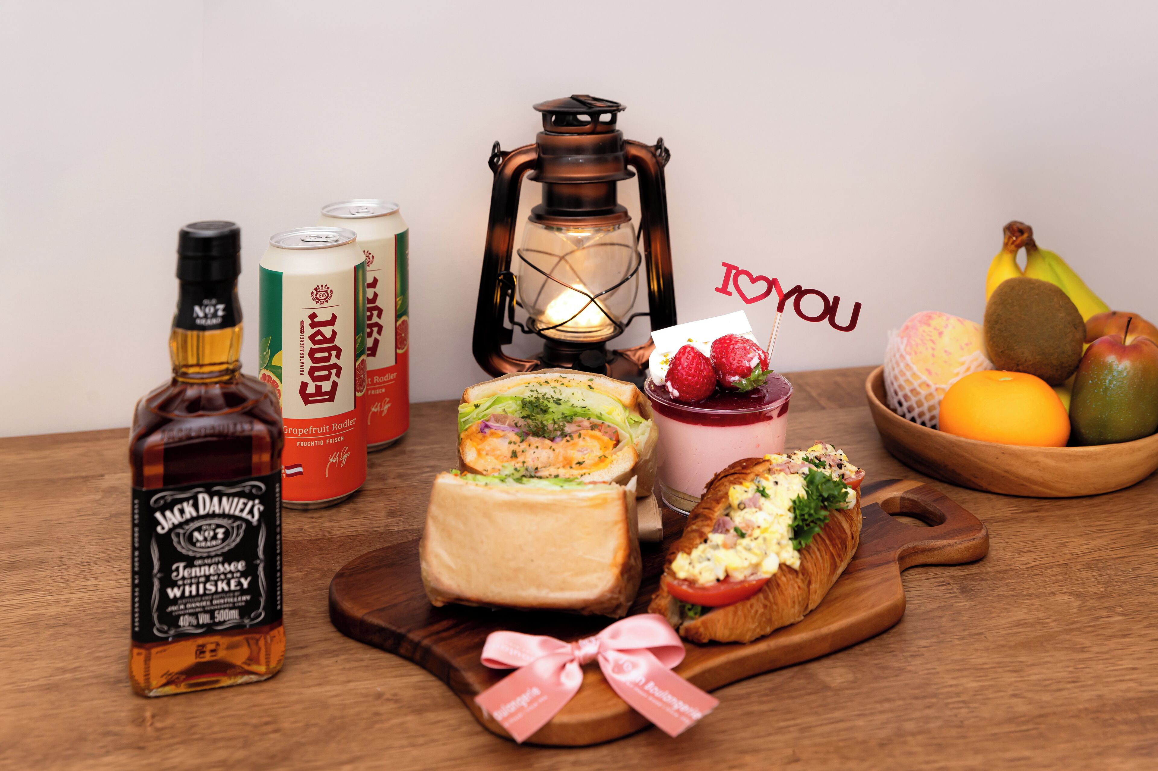 A wooden table with a bottle of Jack Daniel's whiskey, two cans of beer, a sandwich, a dessert, a lantern, a fruit bowl, and a card with 'I ❤️ YOU' written on it.