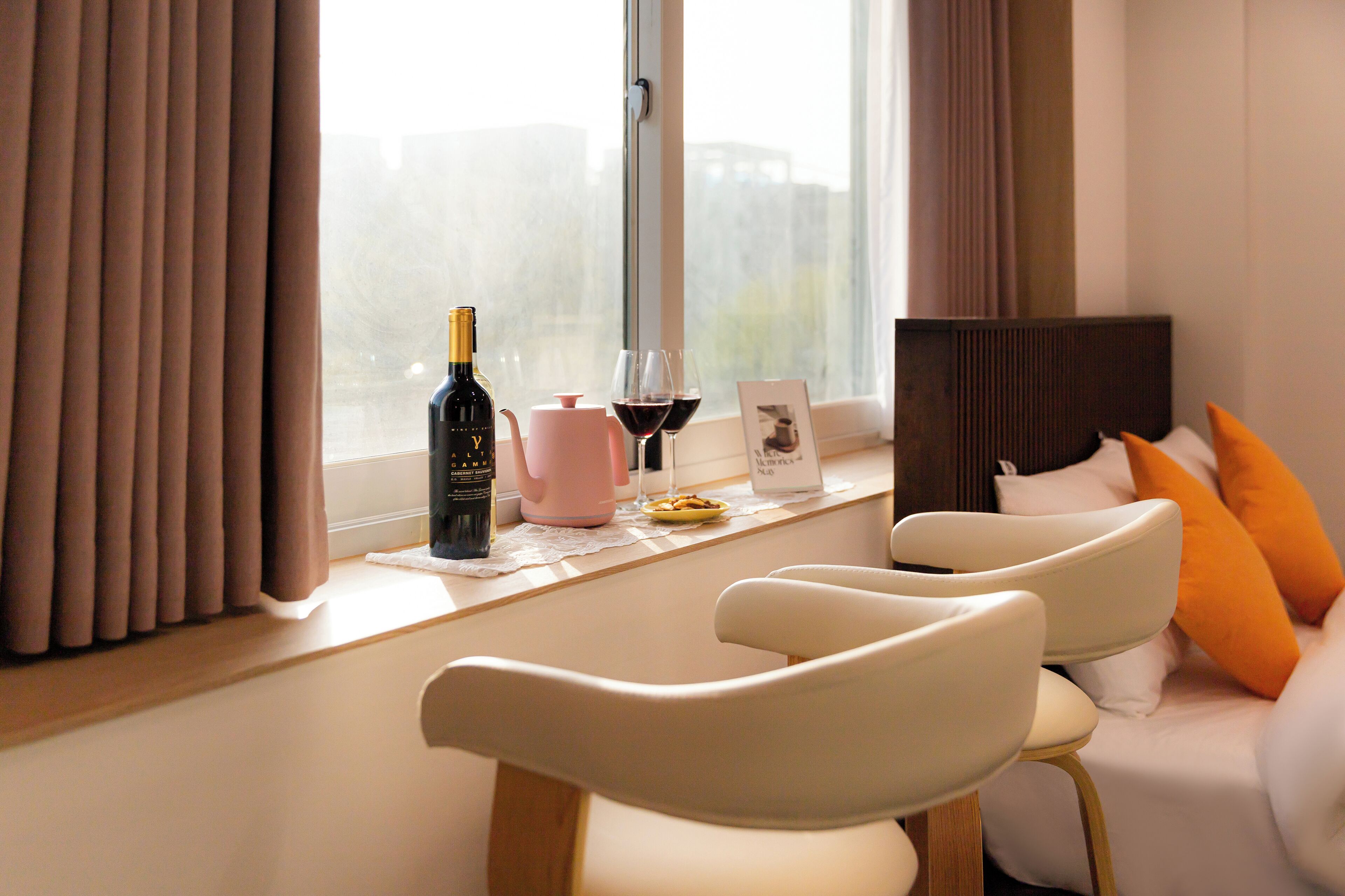 Quarto de hotel moderno com vista para a paisagem urbana, área de jantar com mesa posta para dois e sofá com almofadas laranja.