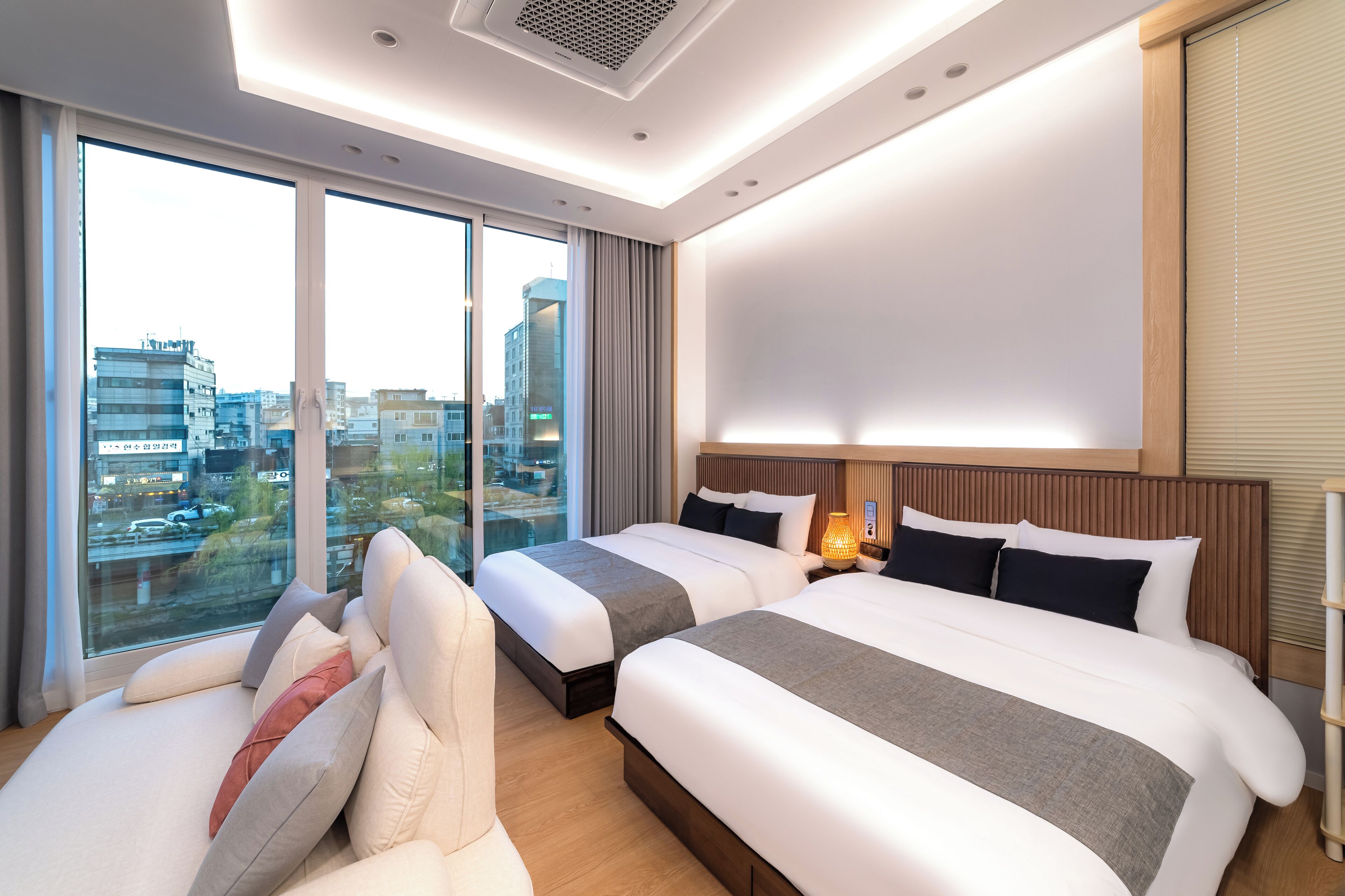 A hotel room with two beds, a large window with a city view, and a modern ceiling design.