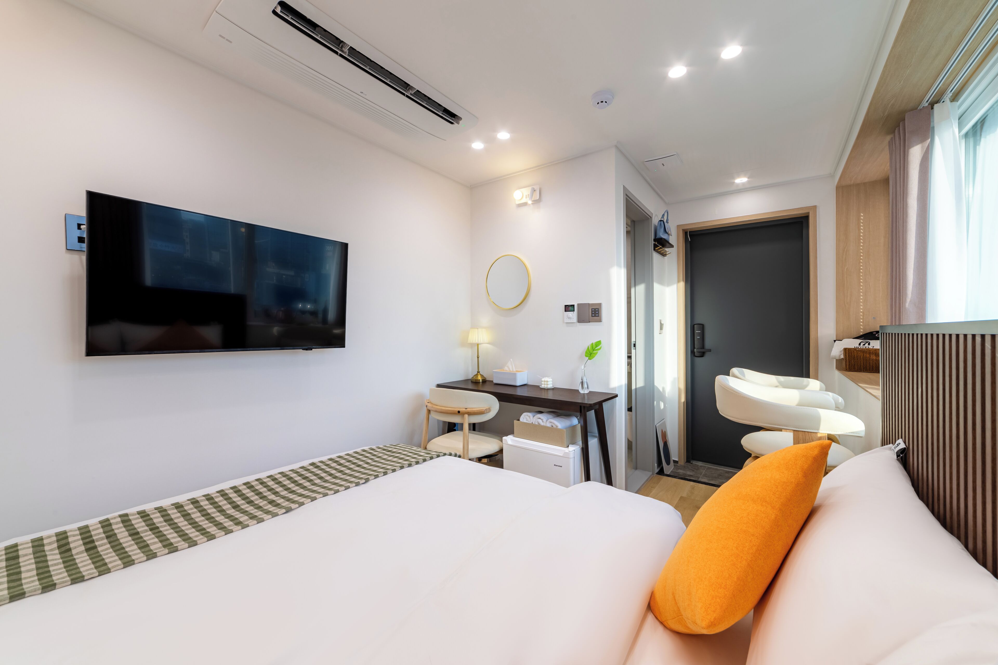 A modern hotel room with a bed, a desk, a chair, a TV, a mirror, and a bathroom.