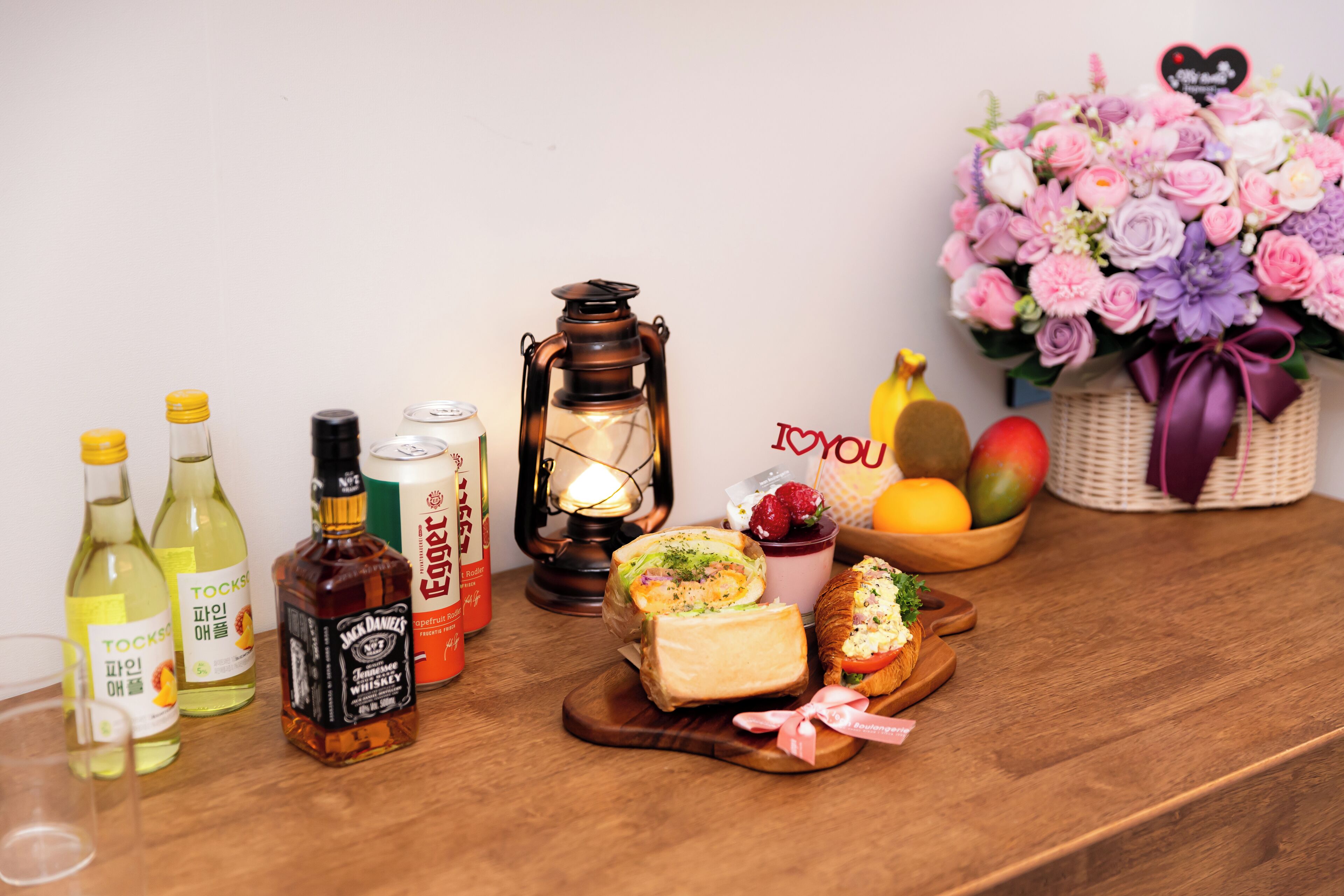A wooden table with various items including bottles, a lantern, sandwiches, fruits, and a floral arrangement.