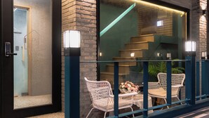 Exterior moderno de un hotel con puerta de vidrio, zona de estar al aire libre con sillas de mimbre y una mesa, y una escalera visible a través del ventanal.