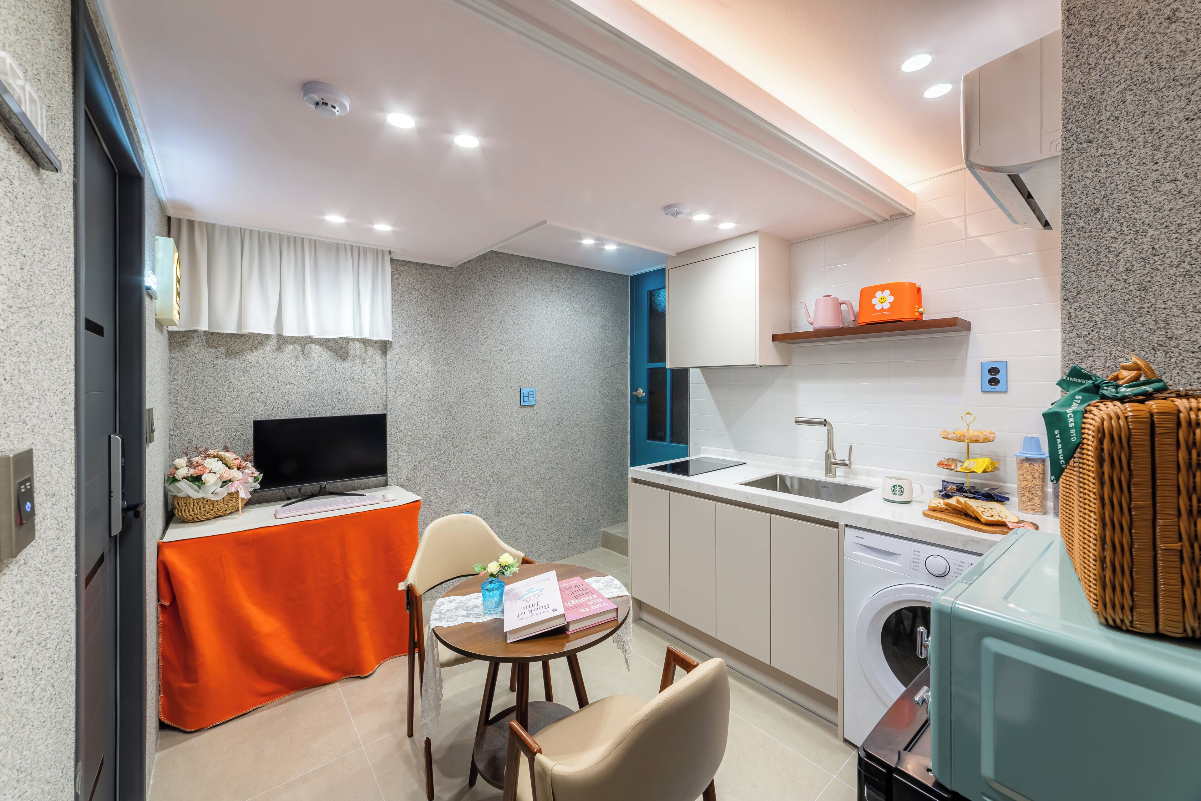 A compact kitchen with a washing machine, a dining area with a table and chairs, and a television on a stand.
