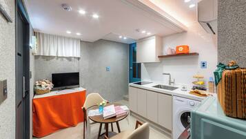 A compact kitchen with a washing machine, a dining area with a table and chairs, and a television on a stand.