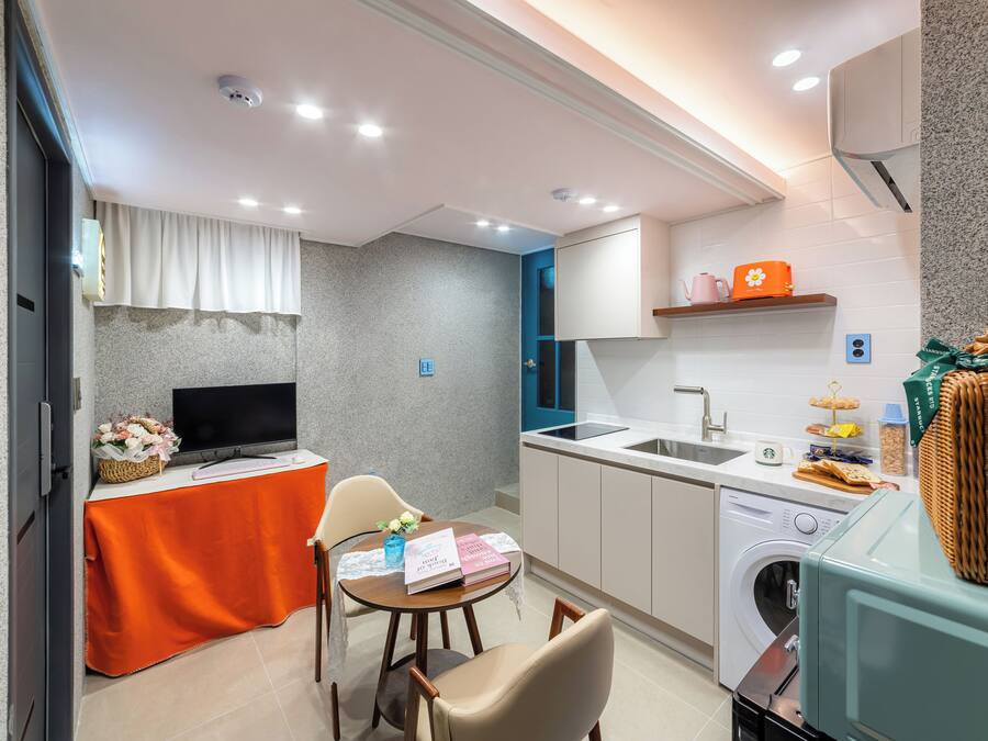 A compact kitchen with a washing machine, a dining area with a table and chairs, and a television on a stand.