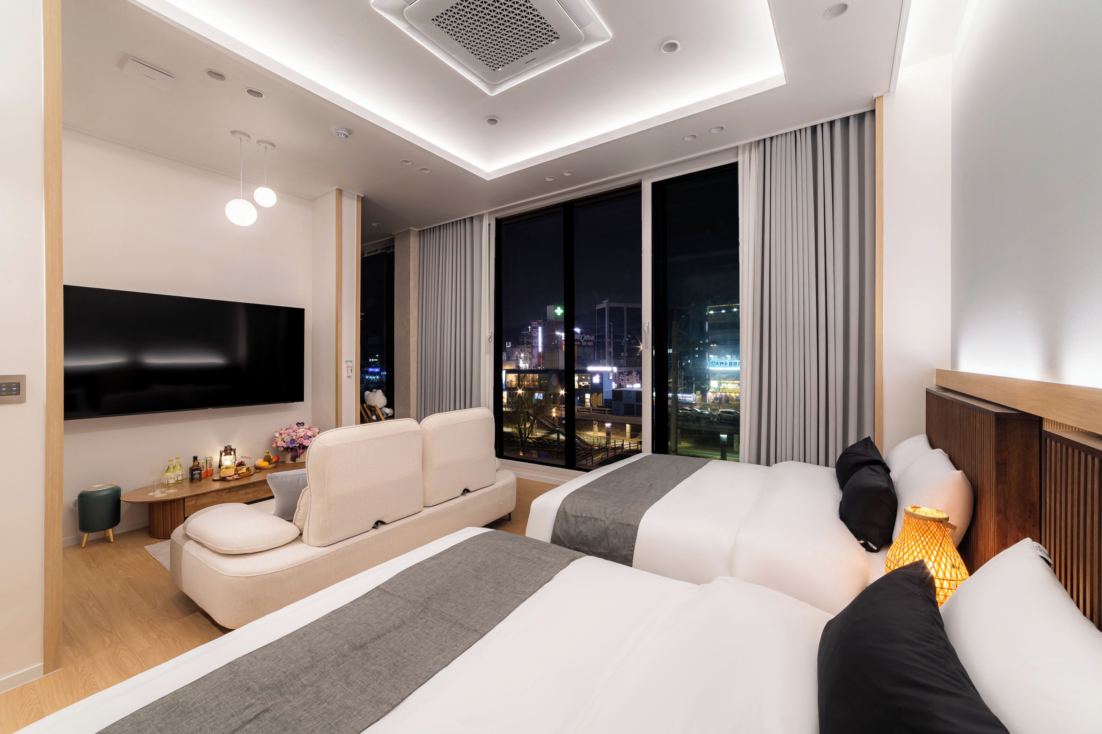 A modern hotel room with a large TV, two beds, a sofa, a small table with drinks, and a city view.