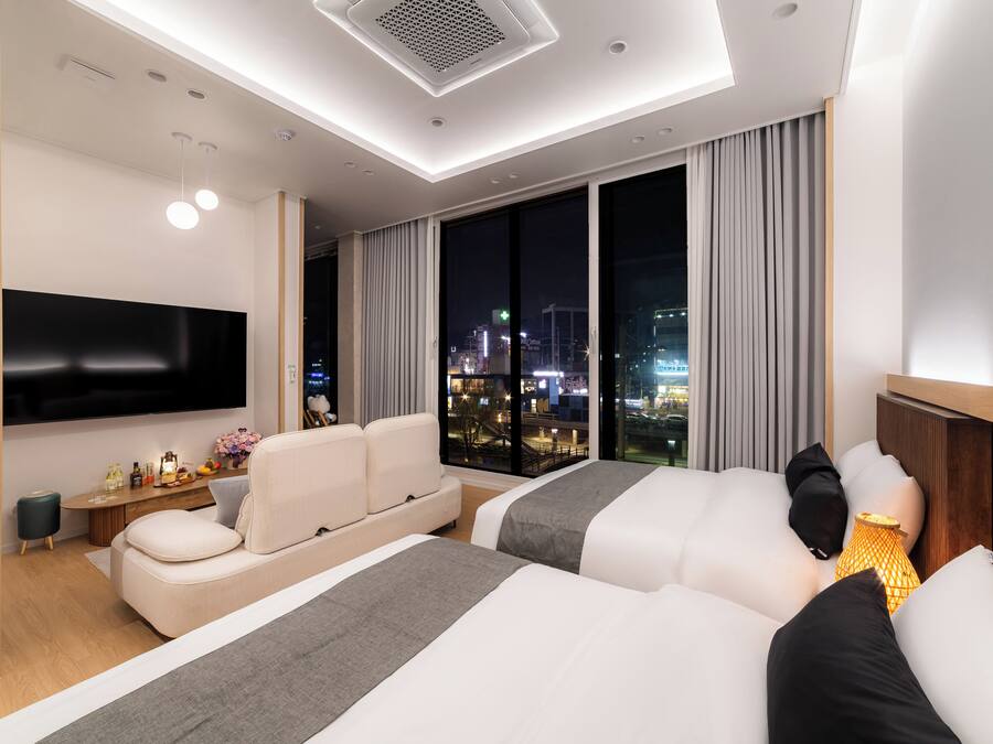 A modern hotel room with a large TV, two beds, a sofa, a small table with drinks, and a city view.