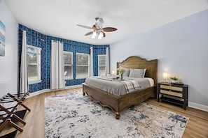 A bedroom with a wooden bed, a ceiling fan, a nightstand with a lamp, and a large window with curtains.