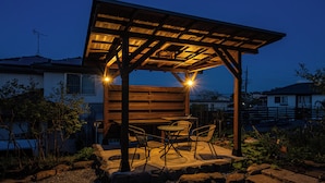Une pergola en bois avec une table et des chaises, entourée d’un jardin agrémenté de rochers et de plantes.