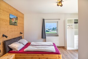 Ein Schlafzimmer mit einem hölzernen Kopfteil, einem Bett mit gemusterten Kissen, einer roten Bettdecke, einem Fenster mit Blick auf Häuser und Berge sowie einem weißen Kleiderschrank.