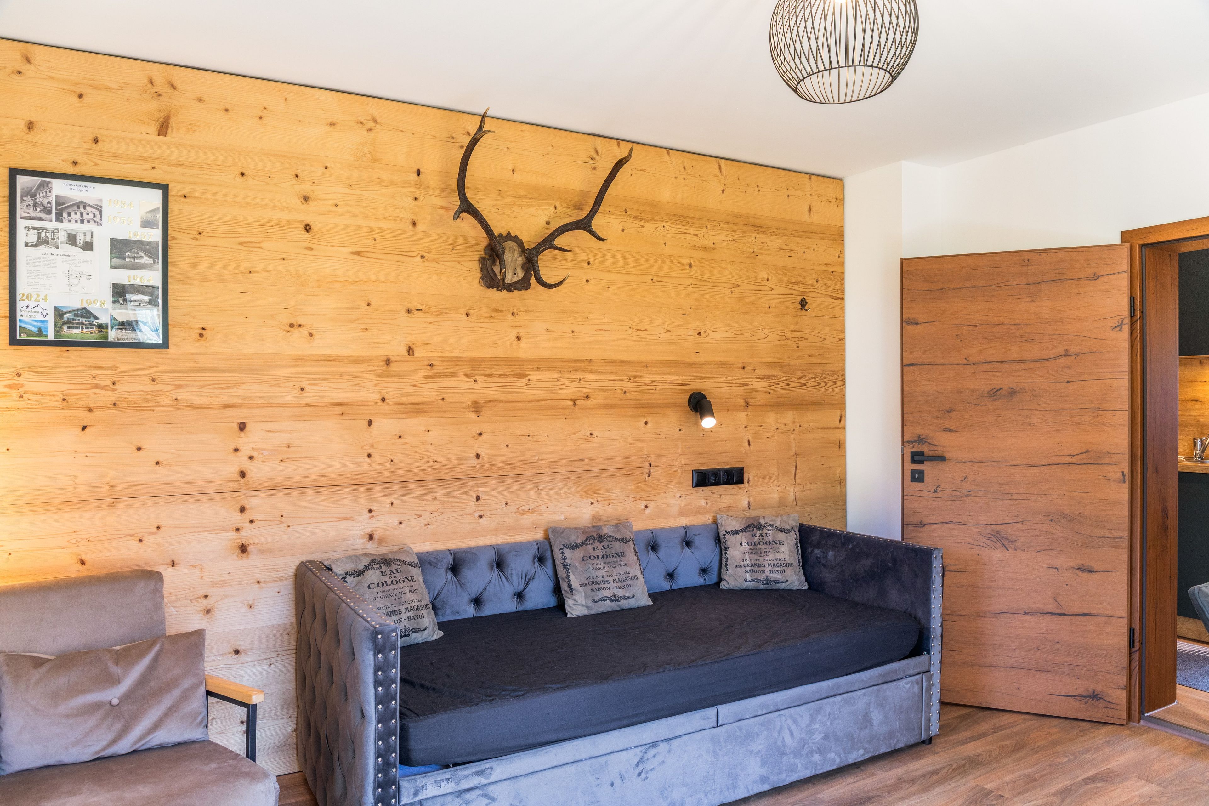 A modern interior with a wooden wall, a mounted antler, a framed photo collage, a sofa with decorative pillows, and a door.
