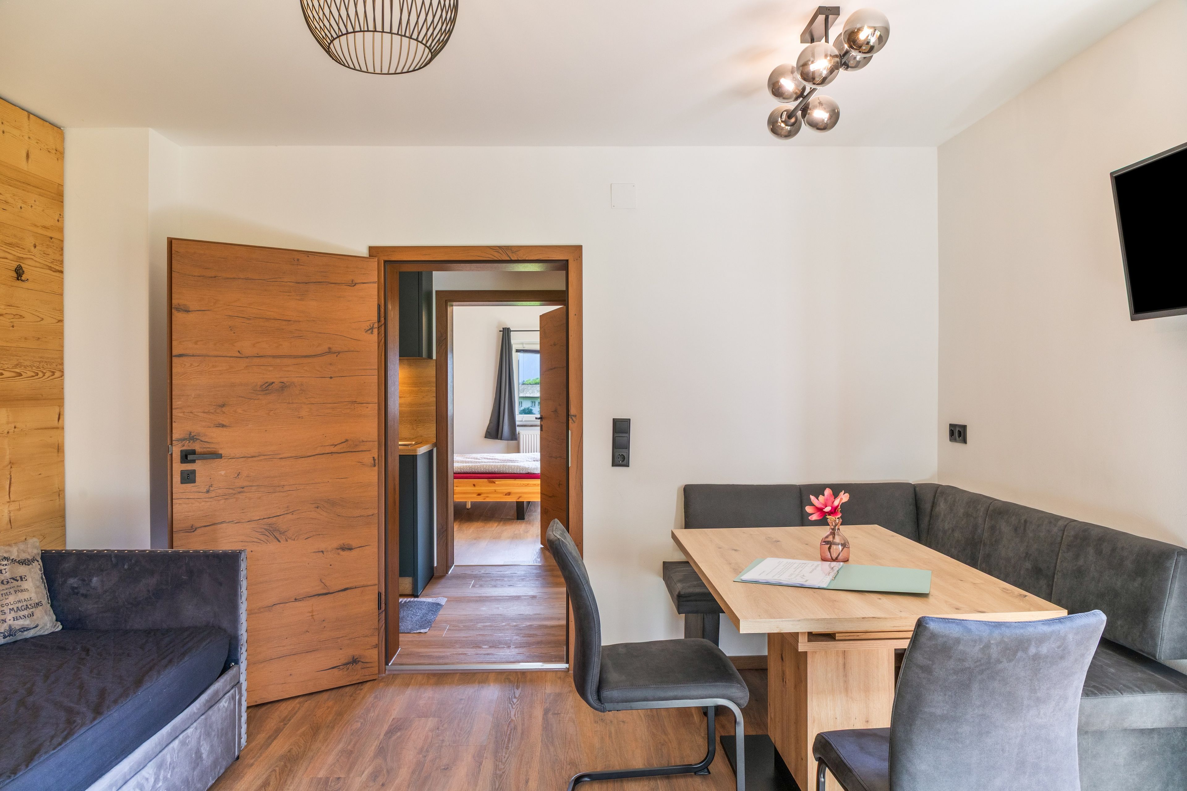 Un intérieur moderne avec une porte en bois, un coin repas comprenant une table et des chaises, un canapé gris et un téléviseur fixé au mur.