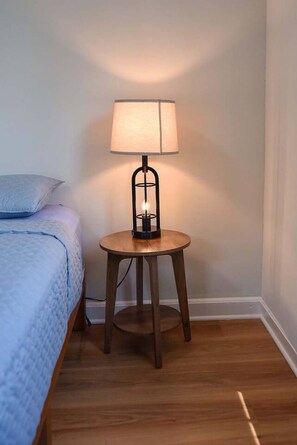 Un comodino piccolo e rotondo in legno con una lampada, accanto a un letto con copripiumino e cuscini blu.