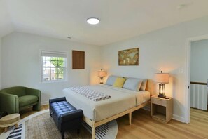 A bedroom with a bed, bedside tables, a chair, a bench, a window, and a painting on the wall.