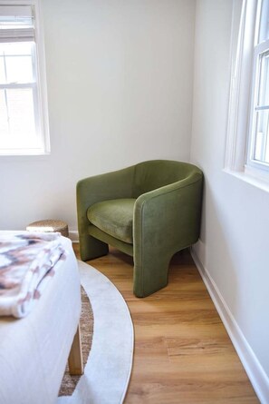 Una camera d'angolo con una poltrona verde, un tappeto rotondo bianco e un letto con copriletto floreale.