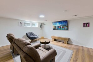 A modern living room with a sofa, a rocking chair, a coffee table, a bench, and a flat-screen TV mounted on the wall.