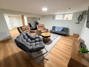 Una sala moderna con un sofá seccional gris, dos sillones marrones, un sillón tipo flexi-bag gris, una mesa de centro de madera y una alfombra gris.