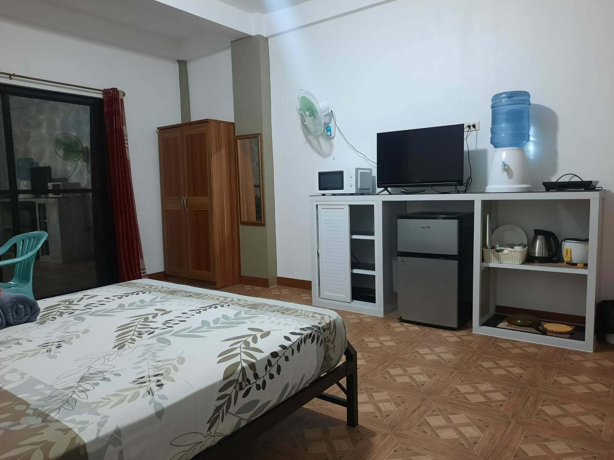 A bedroom with a bed, a wardrobe, a television, a water dispenser, a microwave, a refrigerator, and a small kitchen area.