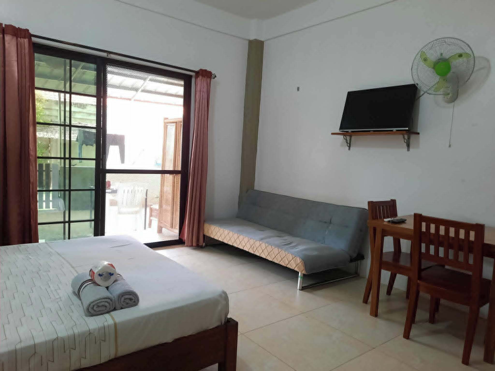A room with a bed, a sofa, a TV, a fan, and a window with curtains.