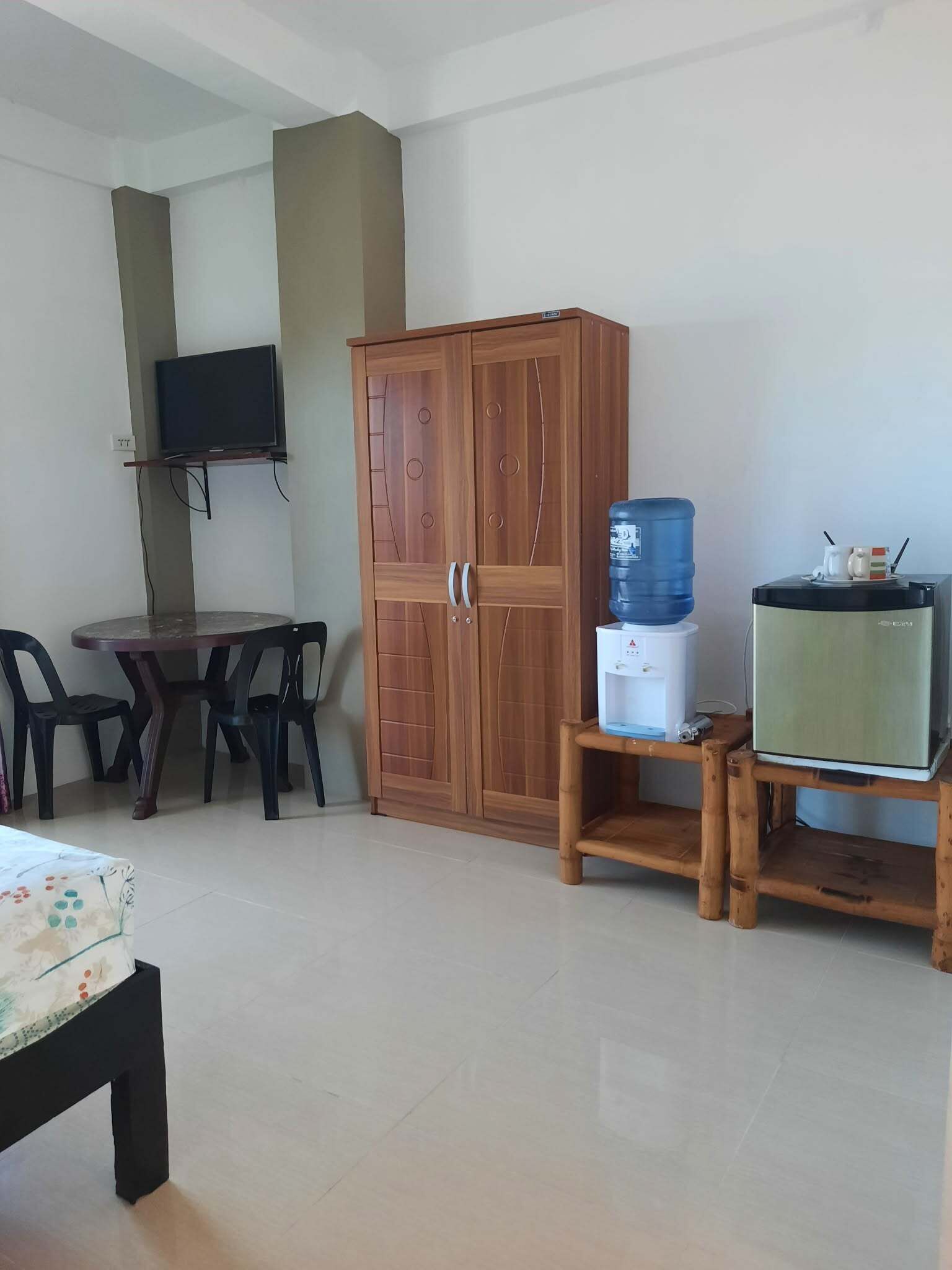 Una habitación con un armario de madera, un televisor montado en la pared, una mesa con sillas, un dispensador de agua y un refrigerador pequeño.