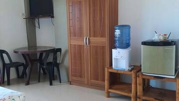 A room with a wooden wardrobe, a television mounted on the wall, a table with chairs, a water dispenser, and a small refrigerator.