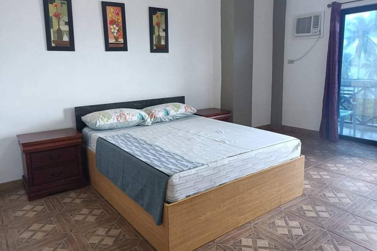Un dormitorio con cama, dos mesitas de noche, aire acondicionado de pared y ventana con cortinas.