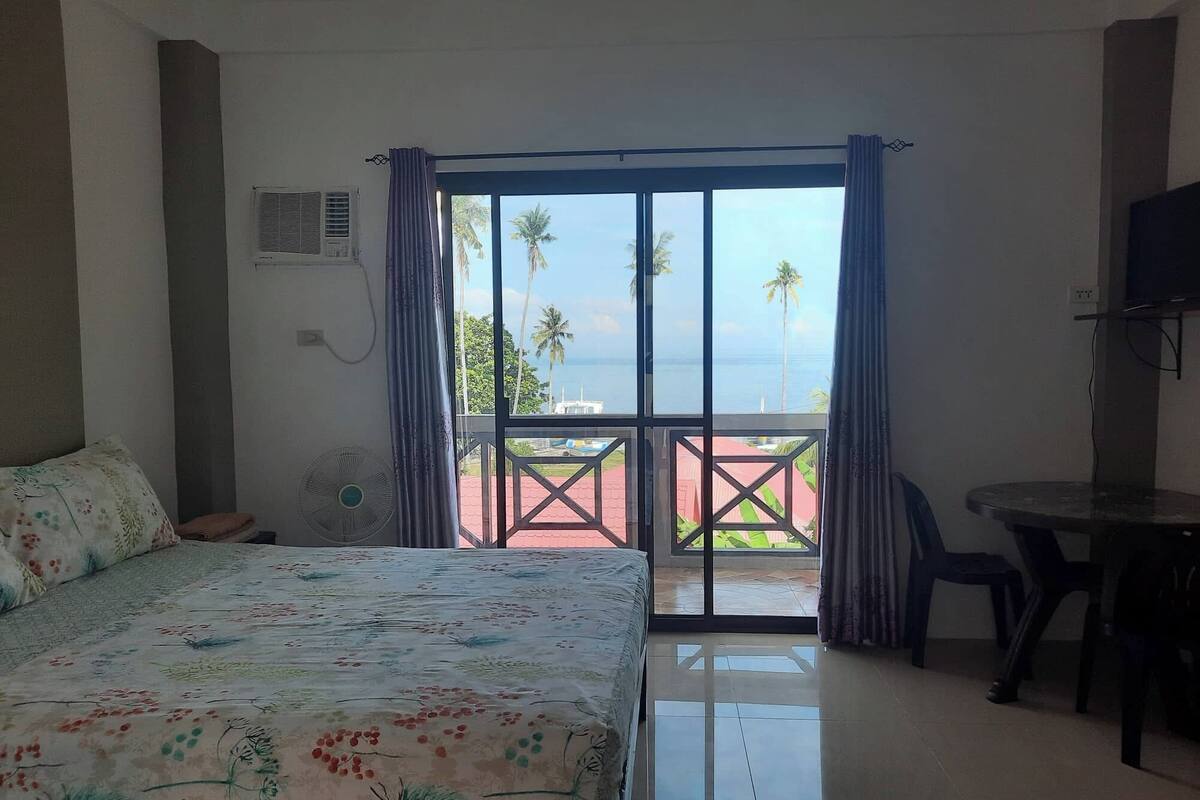 Habitación de hotel con cama, escritorio, silla, televisor y balcón con vista a palmeras y al mar.