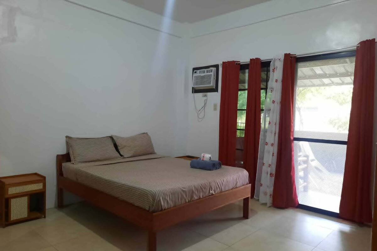 Un dormitorio con cama, una mesita de noche, aire acondicionado instalado en la pared y una ventana con cortinas rojas.