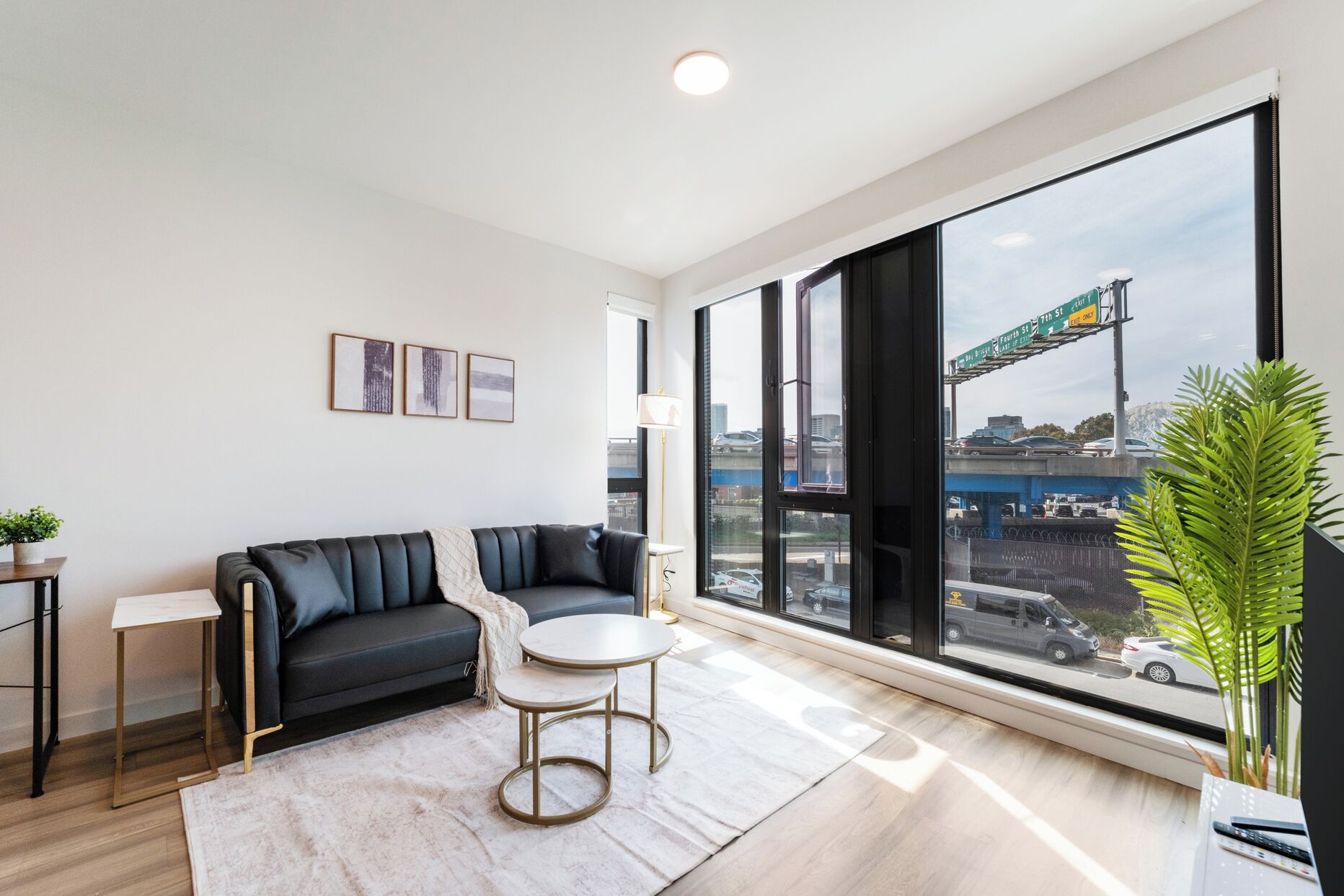 A modern living room with a sofa, a small round table, a potted plant, and large windows offering a view of the city.