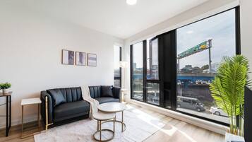 A modern living room with a sofa, a small round table, a potted plant, and large windows offering a view of the city.