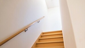 Uma escada com degraus de madeira e corrimão de metal, que leva a um patamar com parede branca e uma luminária no teto.