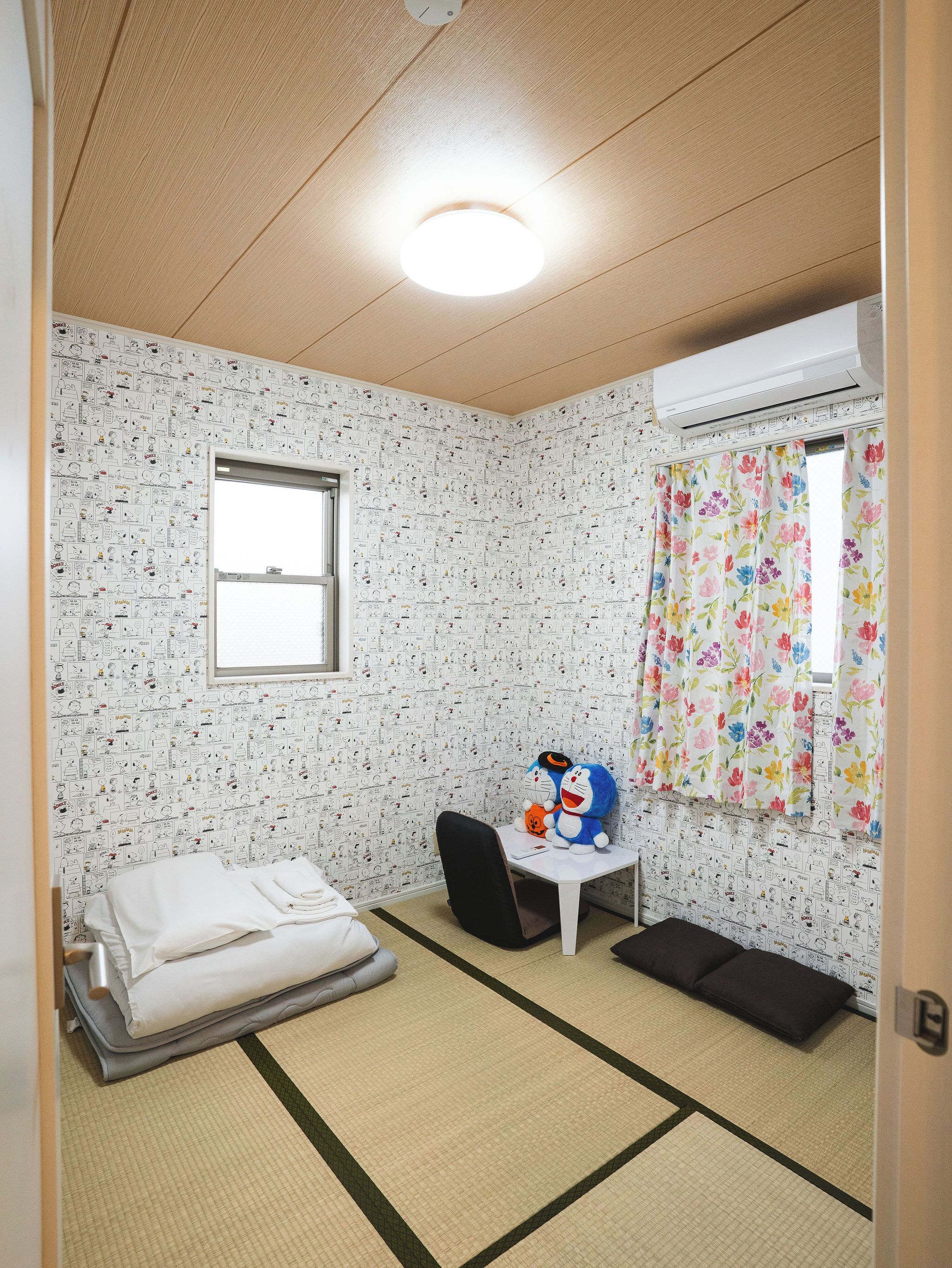 Une pièce avec un sol recouvert de tatamis, un lit, un bureau avec une chaise, une fenêtre avec un rideau et un luminaire au plafond.