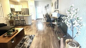 A modern living room with a wooden floor, a coffee table, a TV, a dining area with chairs, and a kitchen in the background. - [HB102] New 3BR near JBSA, Randolph Airforce Base I Fully Stocked + Parking (San Antonio)