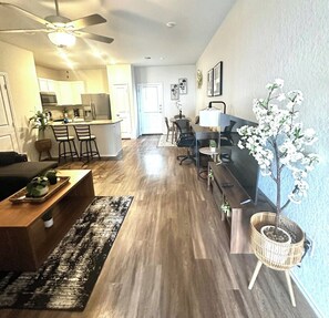 Una sala de estar moderna con piso de madera, una mesa de centro, un televisor, una zona de comedor con sillas y una cocina al fondo.