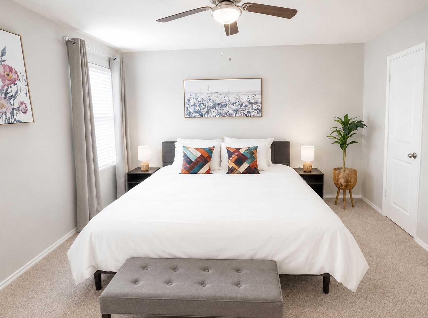 Um quarto com uma cama, duas mesas de cabeceira, um ventilador de teto, uma janela com cortinas, uma obra de arte na parede e um vaso com planta.
